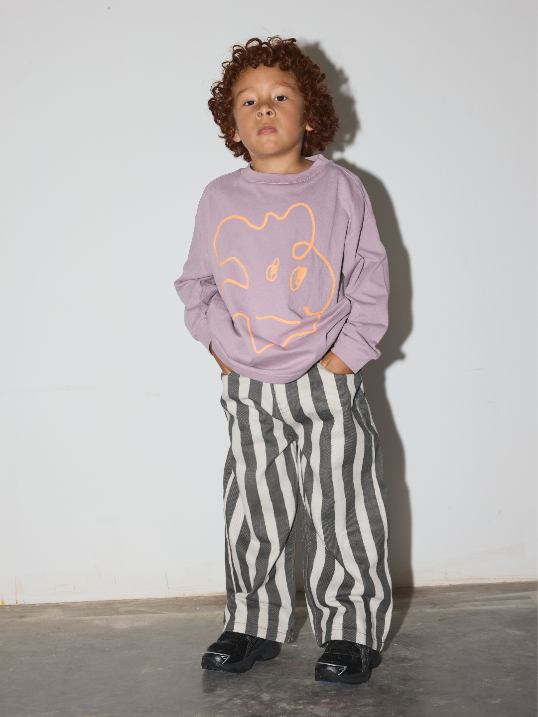 A toddler stands against a plain off-white wall wearing a lavender sweatshirt with a light orange line-art graphic and black-and-white vertically striped jeans.