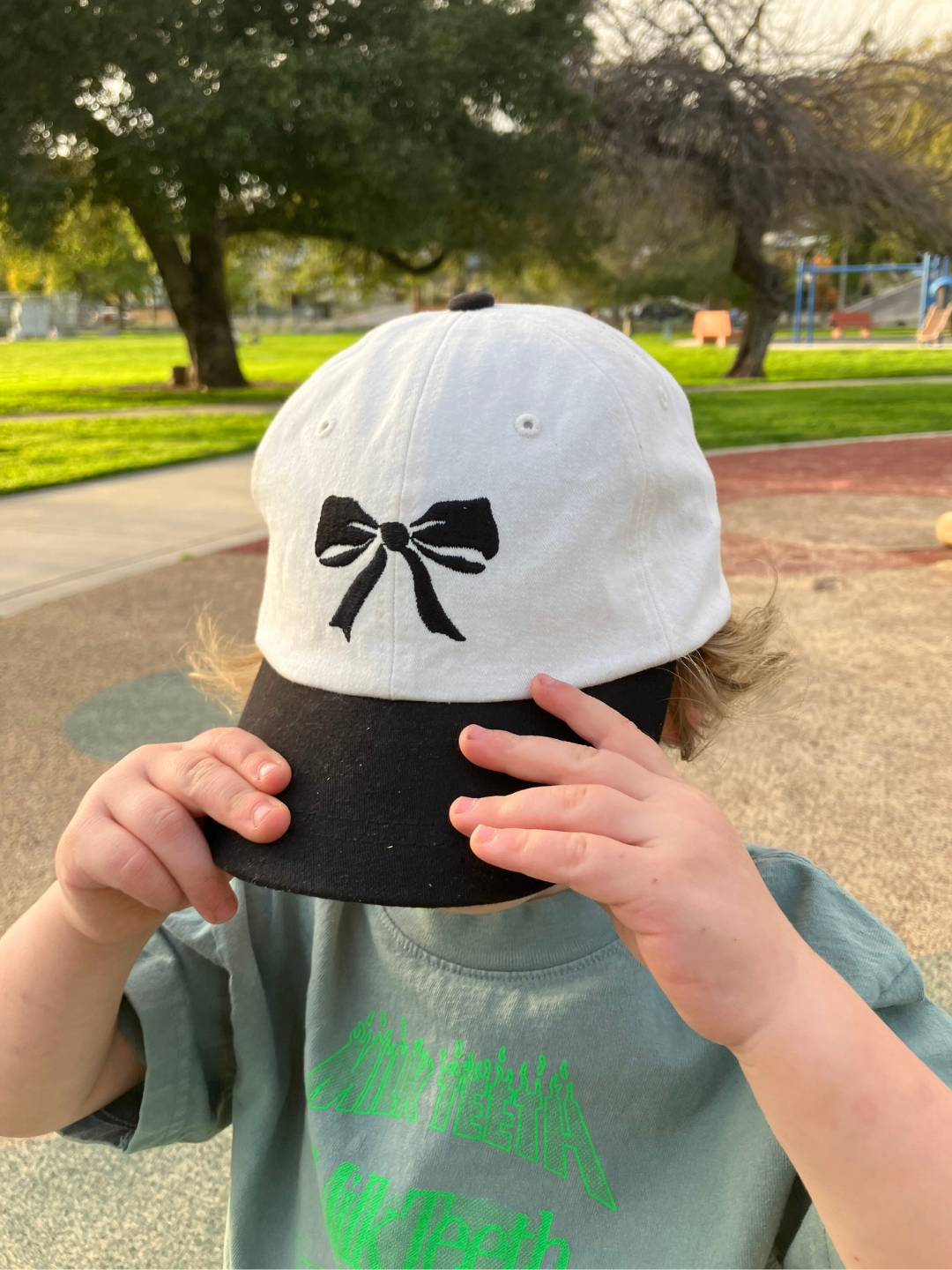 Boy wearing the bow baseball cap in white with a black brim and a black embroidered bow. He cpvers his face with the hat, and wears a green t-shirt with neon green Milk Teeth lettering, outdoors in a playground.