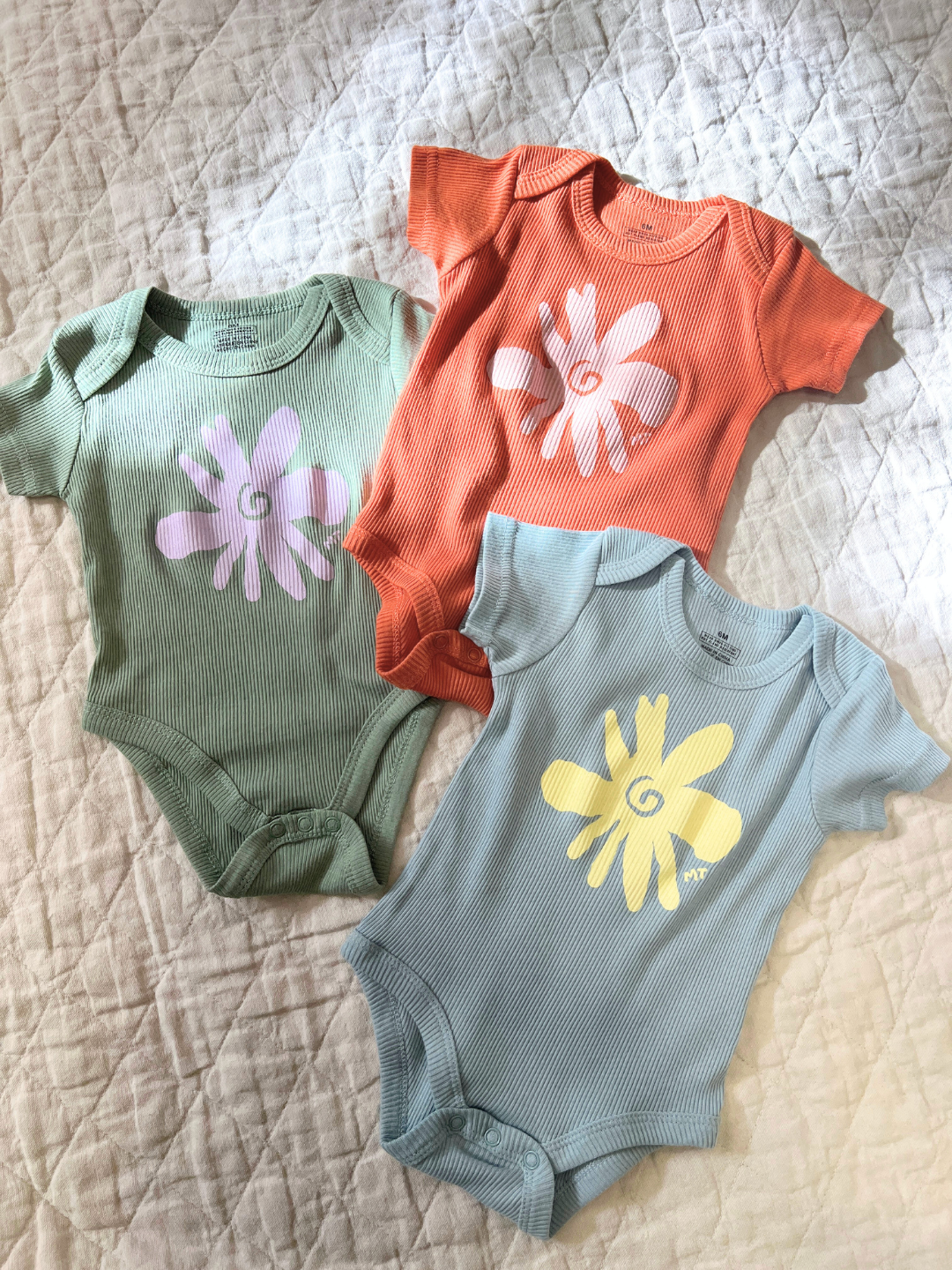 Three RIBBED COTTON ONESIE baby bodysuits with flower prints in green, orange, and blue are displayed on a quilted surface.