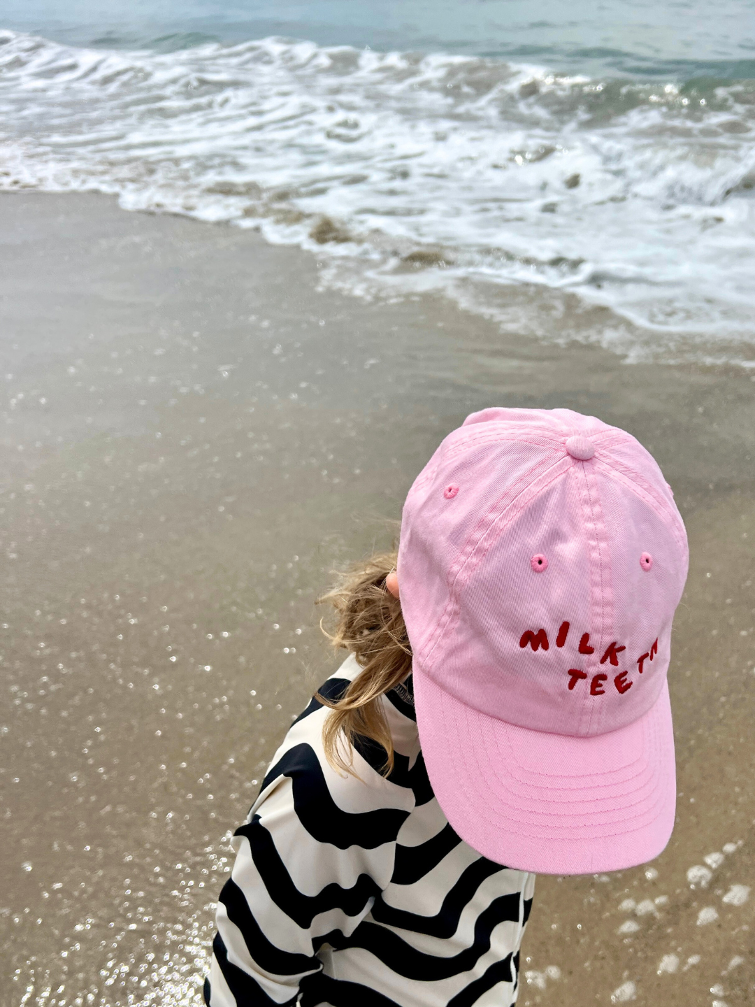 Child wearing the Milk Teeth baseball cap in bubblegum pink with red Milk Teeth lettering, at the beach in a black and white striped swimsuit.