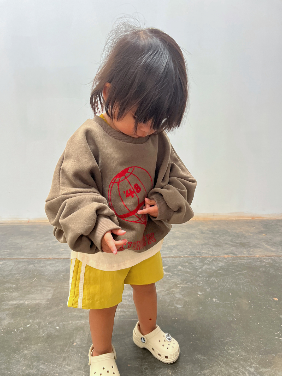 A young child in SPRINTER SHORTS, an oversized brown sweatshirt, and light-colored clogs looks down while standing on a concrete floor against a plain background.