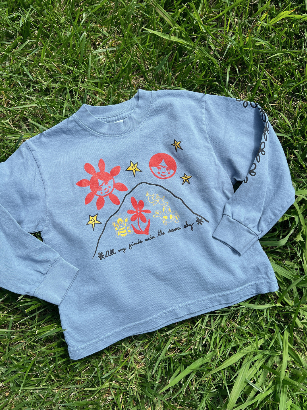 Blue | The ALL MY FRIENDS LONGSLEEVE is a light blue long-sleeve shirt featuring red and yellow graphics of flowers, stars, two faces over a hill, and the text “All my friends under the same sky," shown here laid flat on grass.