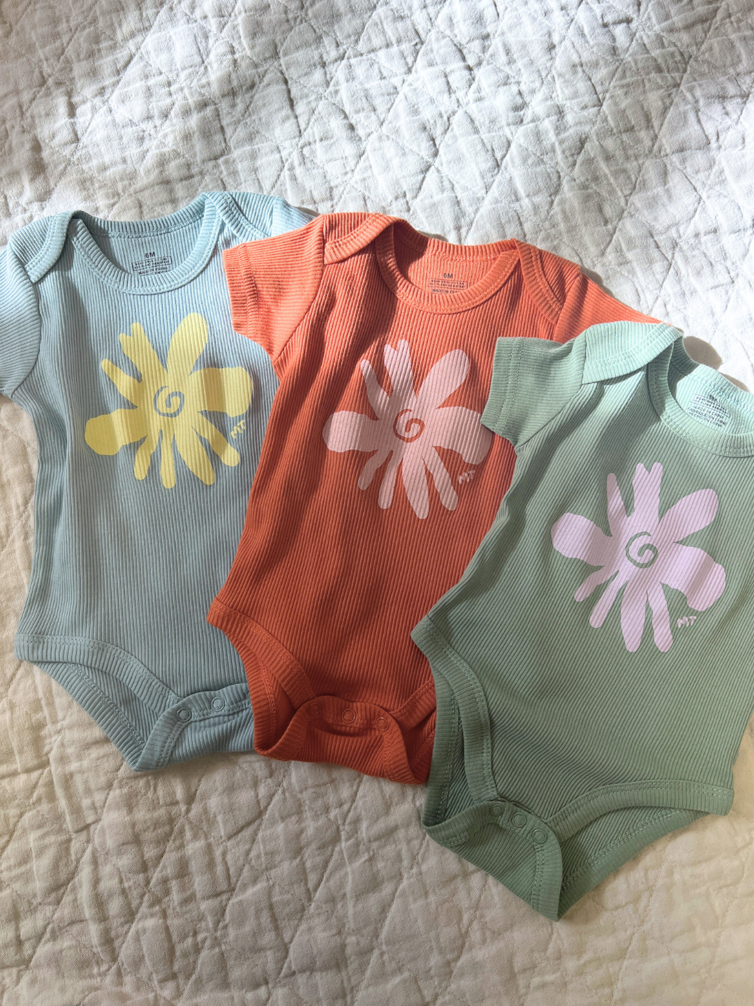 Three RIBBED COTTON ONESIEs featuring delightful flower designs in yellow, orange, and pink are arranged on a quilted surface, perfect for your little one's milk teeth moments.