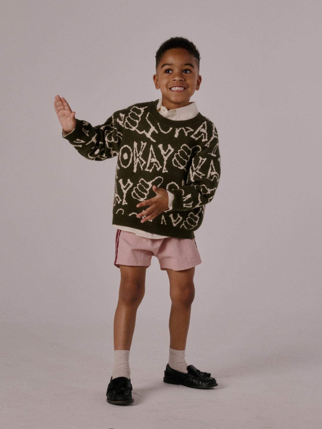 Olive | A young boy stands on one leg, smiling and raising his left hand. He wears the OKAY SWEATER with a green pattern, pink shorts, socks, and black shoes against a simple backdrop.