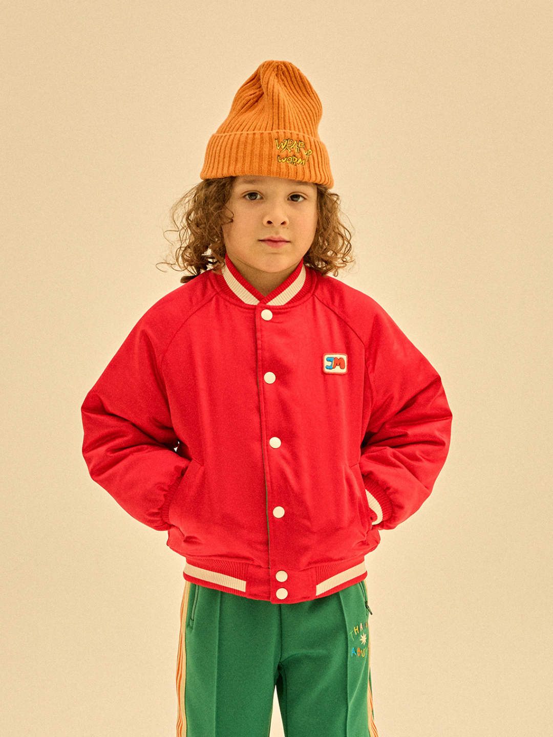 A young child with curly hair wearing an orange knit hat, the BOOM REVERSIBLE JACKET in red with a small patch on the chest, and green pants with colorful stripes stands against a beige background. The child's hands are in the jacket pockets, showcasing ideal merchandising for winter wear.