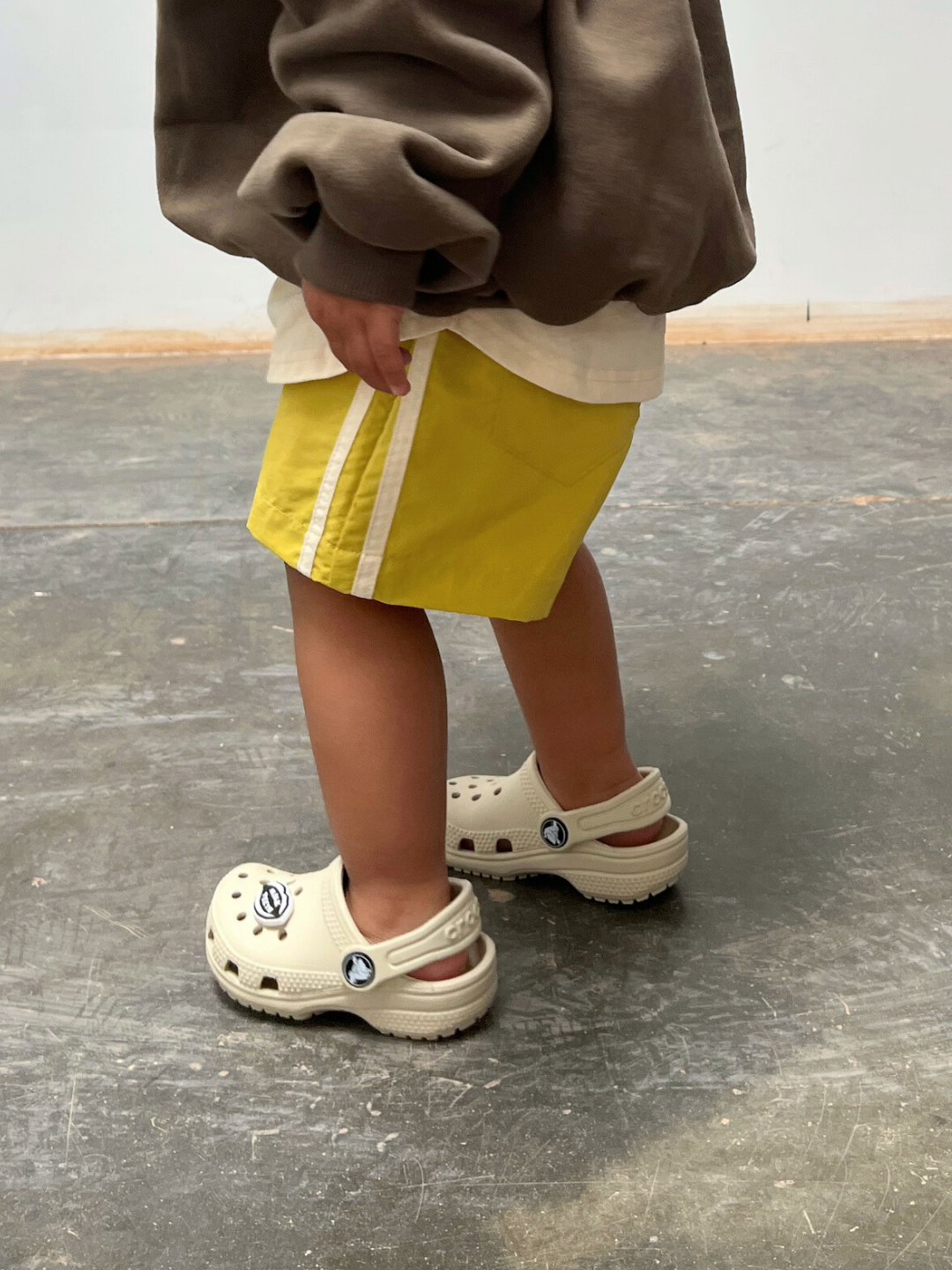 A child wearing a brown sweatshirt, SPRINTER SHORTS in yellow with white stripes, and beige Crocs stands on a concrete floor.