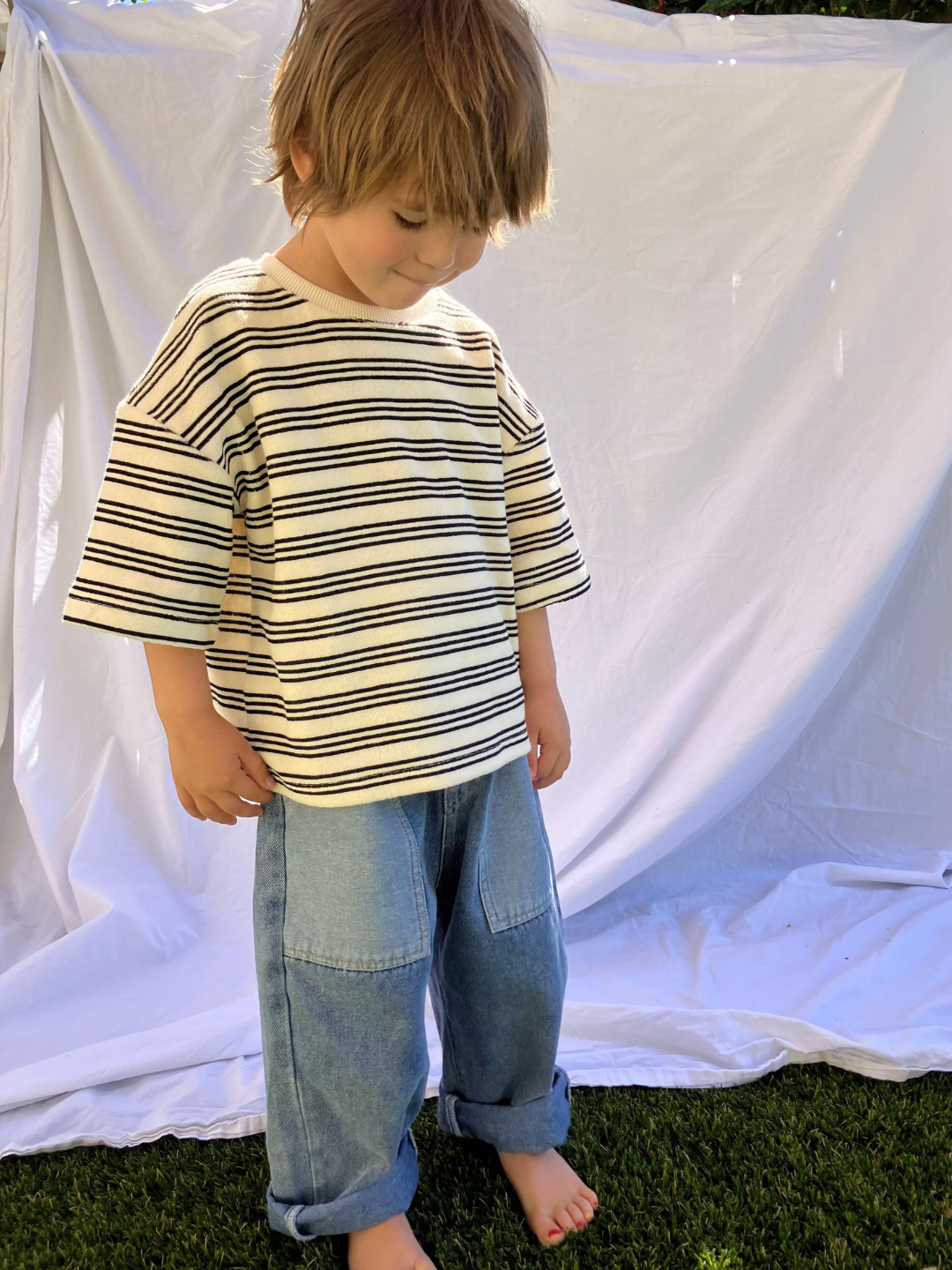 A child wearing the Double Trouble baggy jeans in medium-blue denim with two large patch pockets of lighter denim. He wears an oversized striped t-shirt in cream, with black stripes, and stands in front of a white sheet, standing on grass.