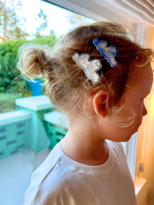 Second image of Two CLOUDS HAIRCLIPS, one in pearlescent white and the other in light blue, rest on a light gray surface with shadows cast beneath them. These hairclips feature alligator clip backings for versatility with various hair types.