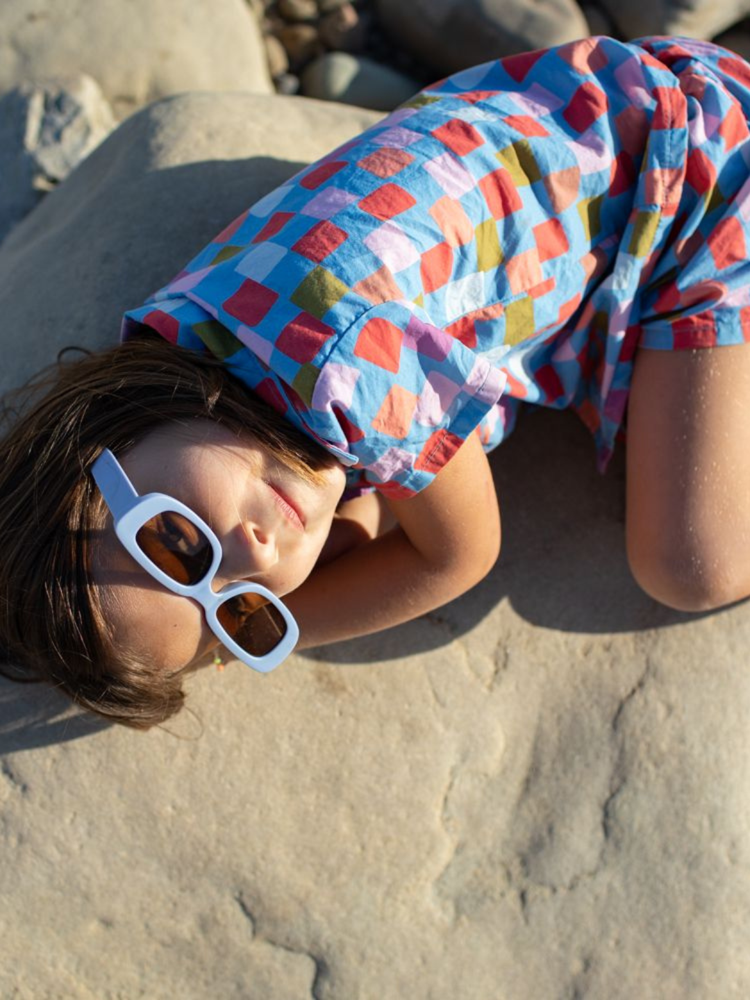A child lying on a rock wearing a kids' shirt and shorts set in a pattern of red, orange, pink, lilac and green squares on a blue background