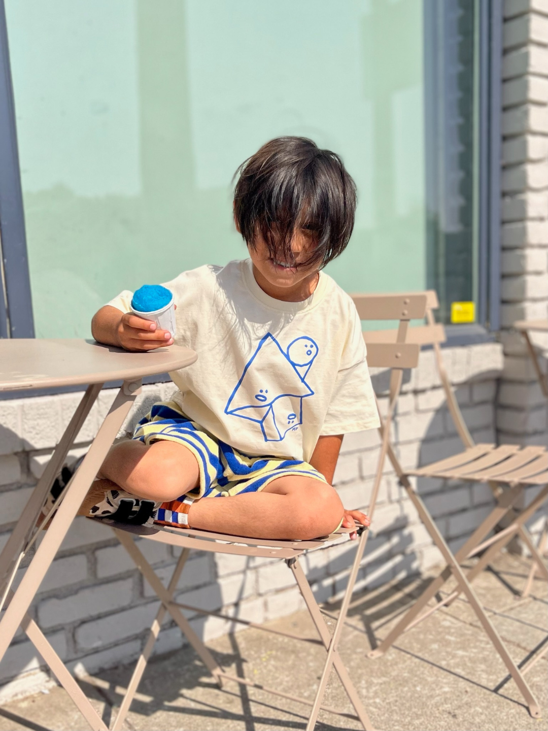 Cream | A kid wearing the Jumble Tee in cream with blue shapes outline with funny faces in the center. He wears striped shorts, is holding a blue sno cone and sits on a cafe chair outdoors.