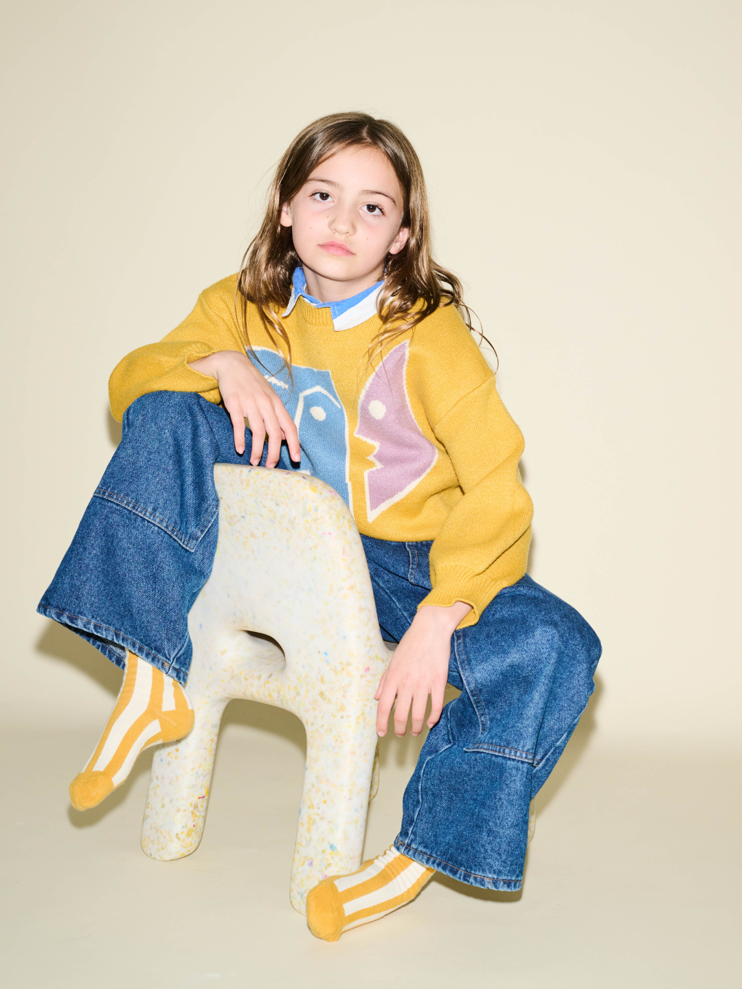 Marigold | A child with long hair sits on a uniquely shaped chair, wearing a comfy yellow sweater with a face design, blue jeans, and the STRIPE SOCK made of stretchy cotton. The background is plain beige.