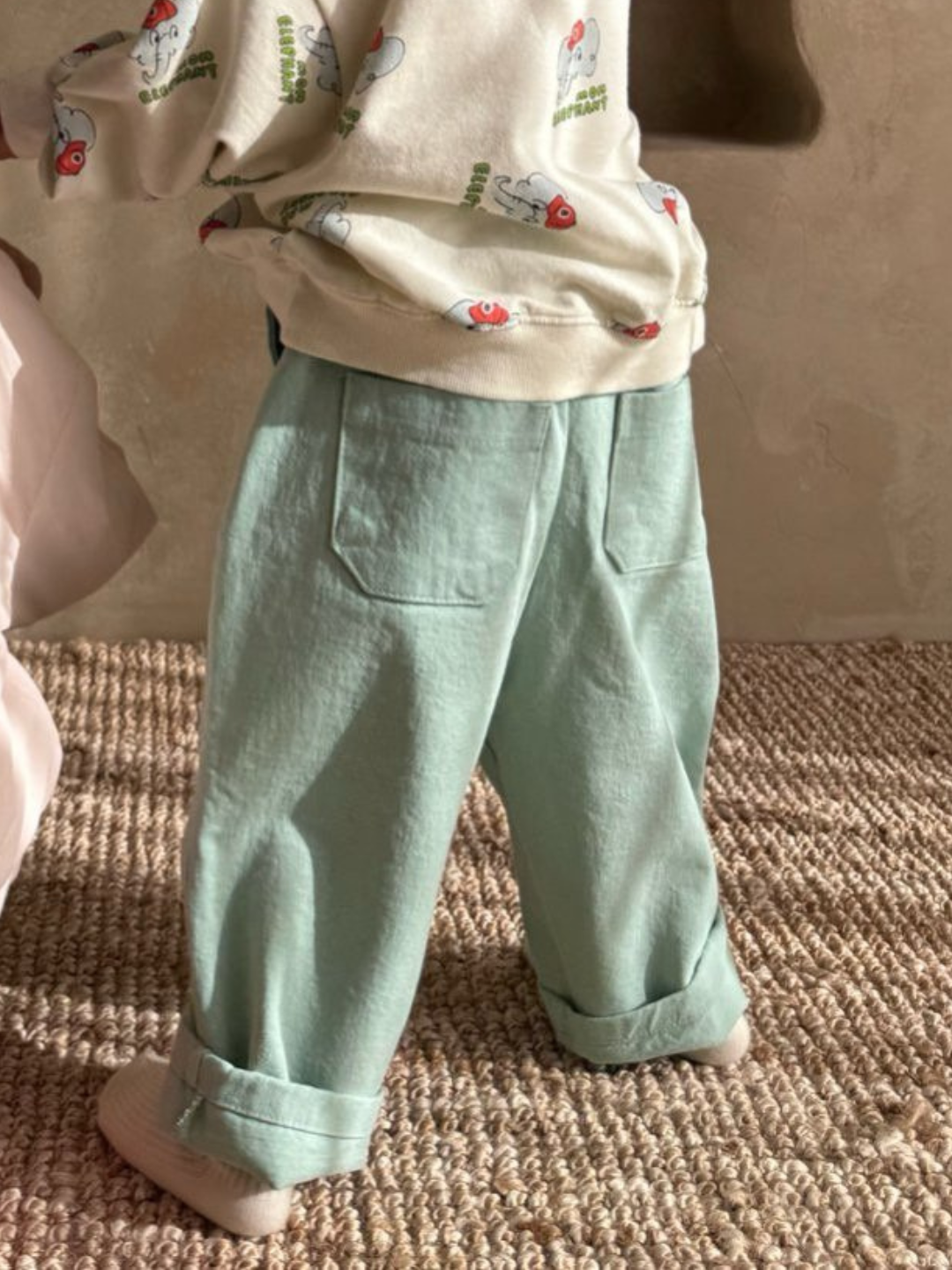 A child stands on a woven rug wearing a patterned top and Tomboy Pants in slouchy mint green from a renowned Korean kids' brand.