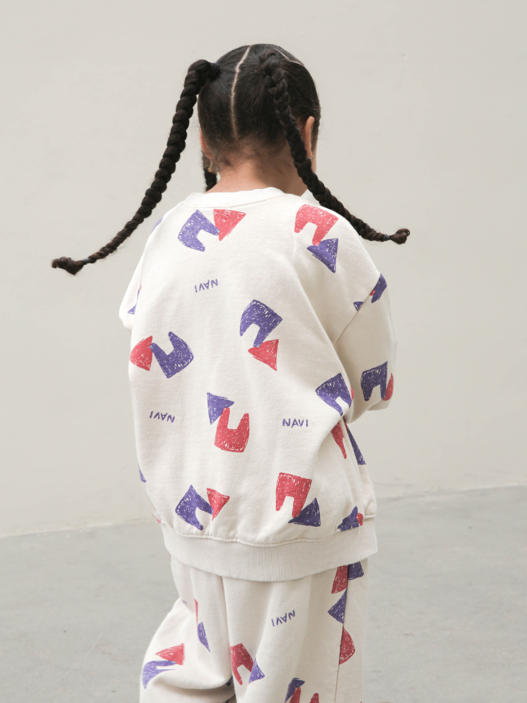Oatmeal | Back view of child wearing beige crewneck sweatshirt with an all over pattern of red and purple shapes and the brand name Navi, with matching sweatpants. She stands on a beige background and wears her hair in three long braids.