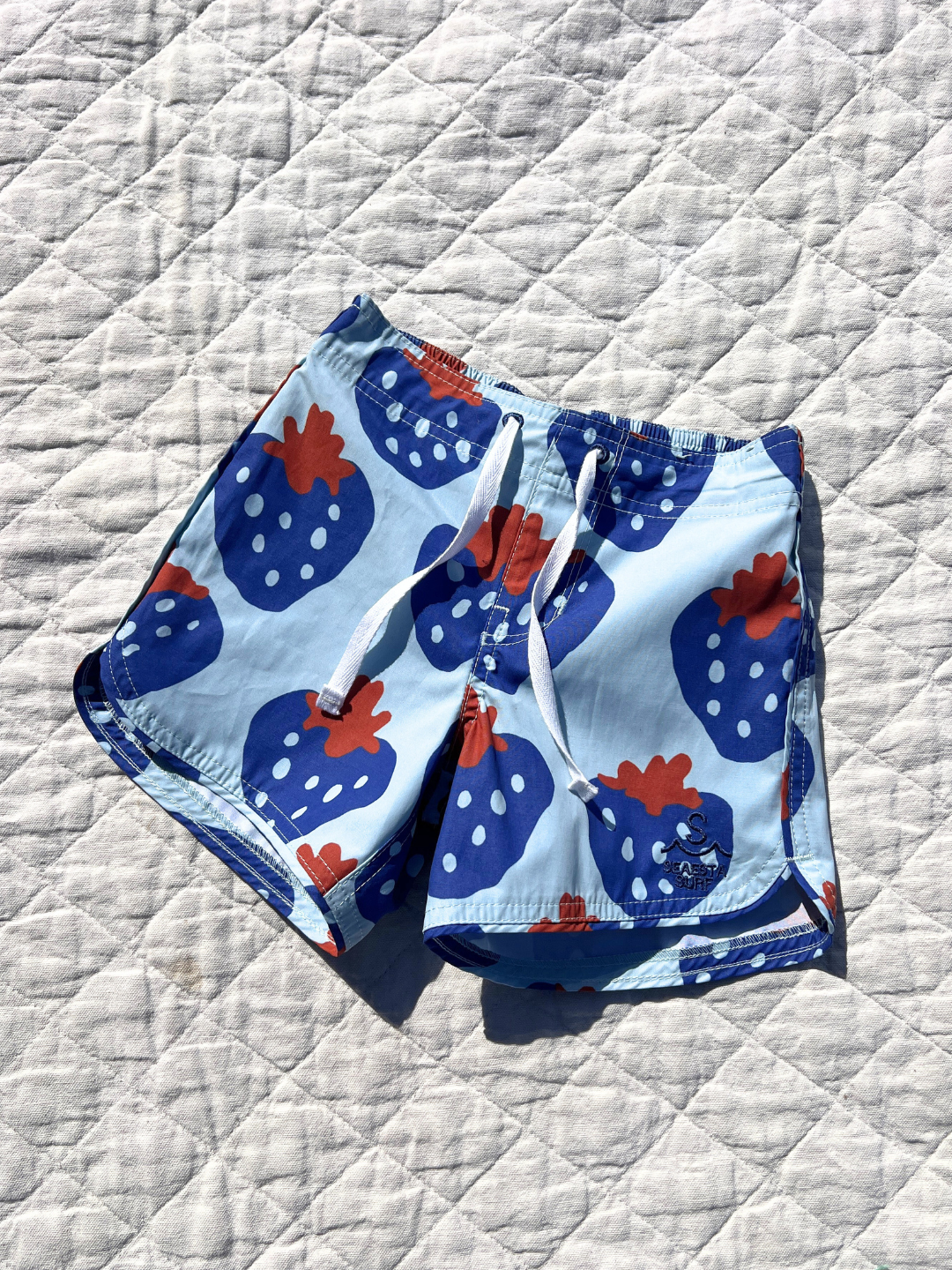 STRAWBERRY BOARDSHORTS: These playful swim trunks feature a fun strawberry pattern with red leaves and white dots on a quilted white background. Made from sustainable materials, they offer a perfect balance of style and eco-friendliness.