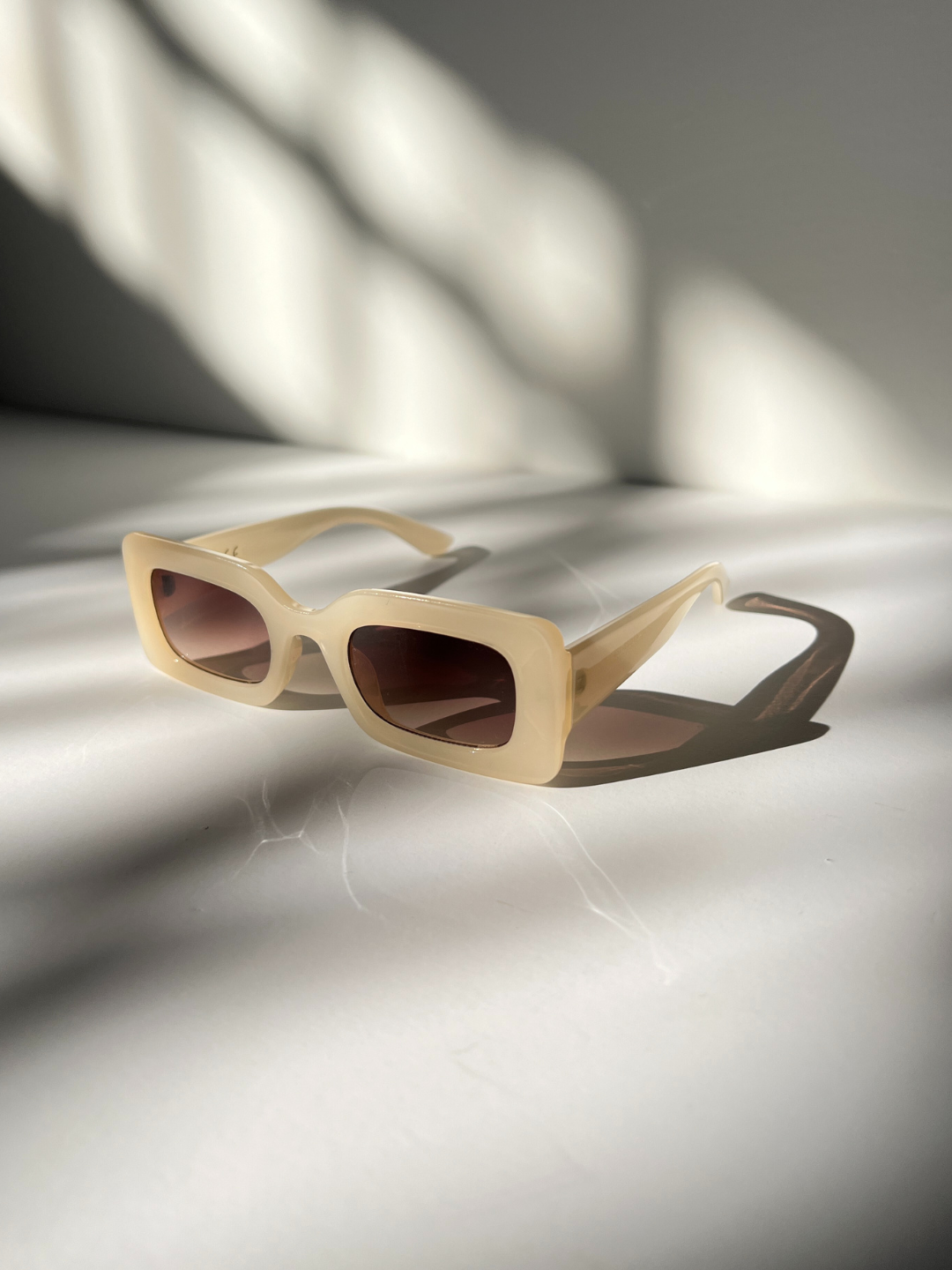 Kids rectangle sunglasses in cream, on a grey background with areas of sunlight, creating shadows of the sunglasses.
