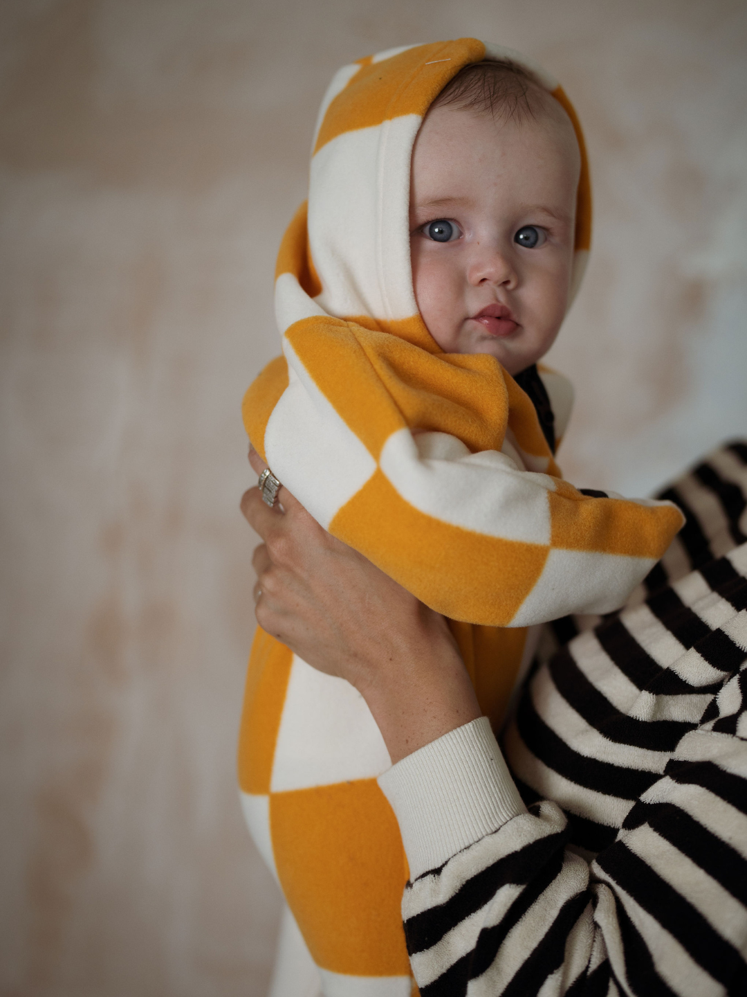 A baby wearing a hooded stroller suit in a bold apricot and white checkerboard pattern 