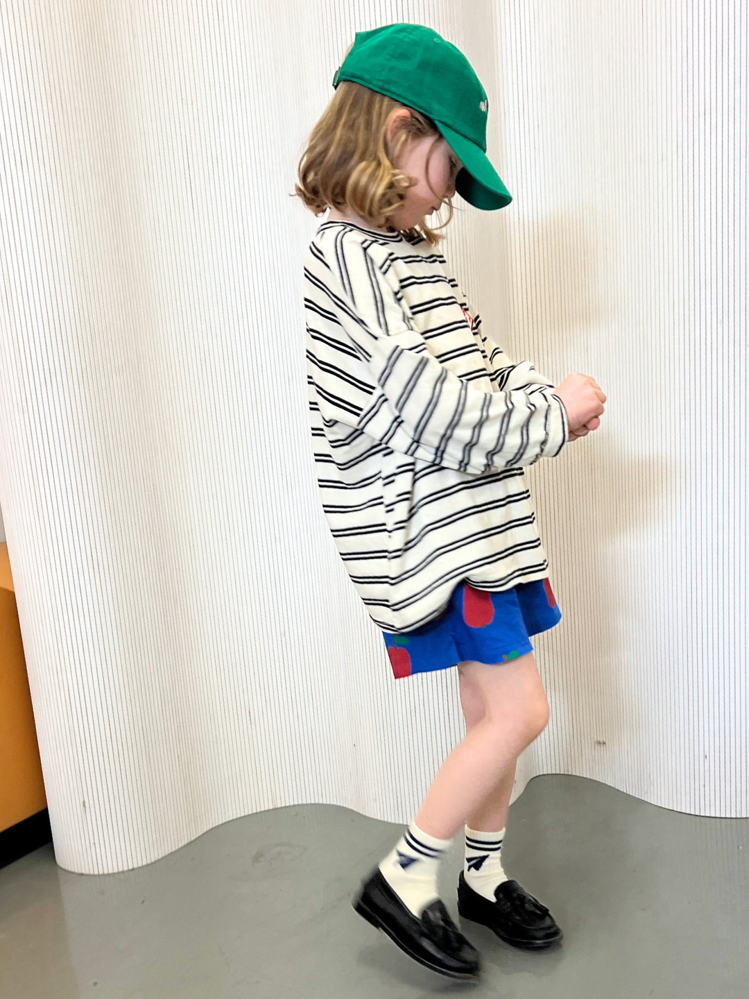 Black & White | A child wearing a green cap, the PEPPER LONGSLEEVE TEE in stripes, and blue shorts stands against a light background, embodying the playful charm of a Korean kids brand.