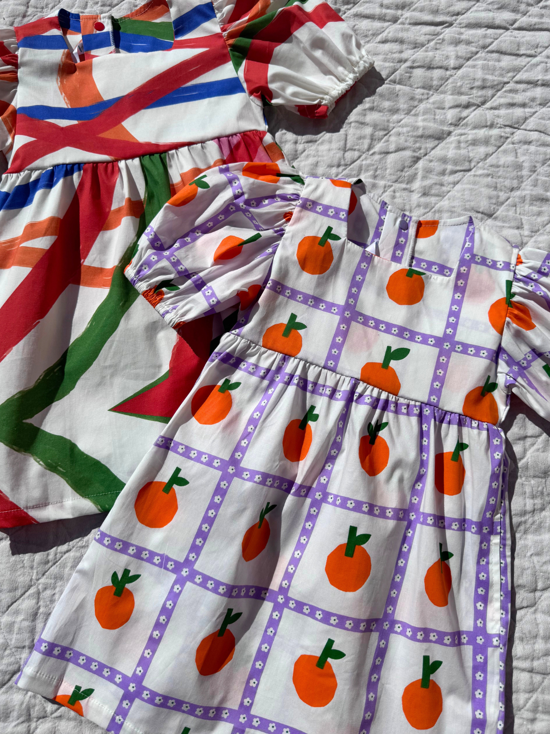 Oranges | A close up of the kid's Glow dress with an all-over oranges print and colors print laid plat on quilt