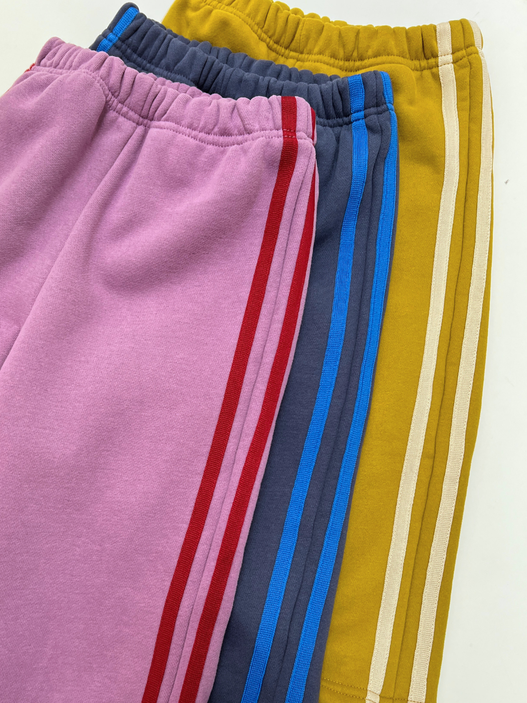 Grape | SUNDAY TRACKPANTS: Three pairs in pink, blue, and mustard yellow with dual side stripes are laid out overlapping on a white surface, offering a cozy, slouchy style perfect for relaxed days.
