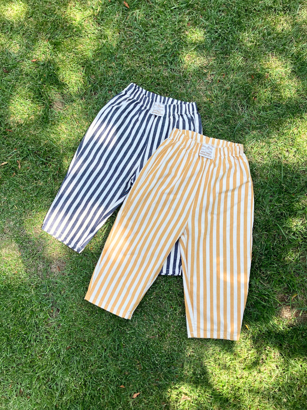 Navy | Two pairs of PONY STRIPE PANTS are laid out on the grass. One pair is black and white, while the other is yellow and white. Both feature an elasticated waistband and a relaxed fit for ultimate comfort.