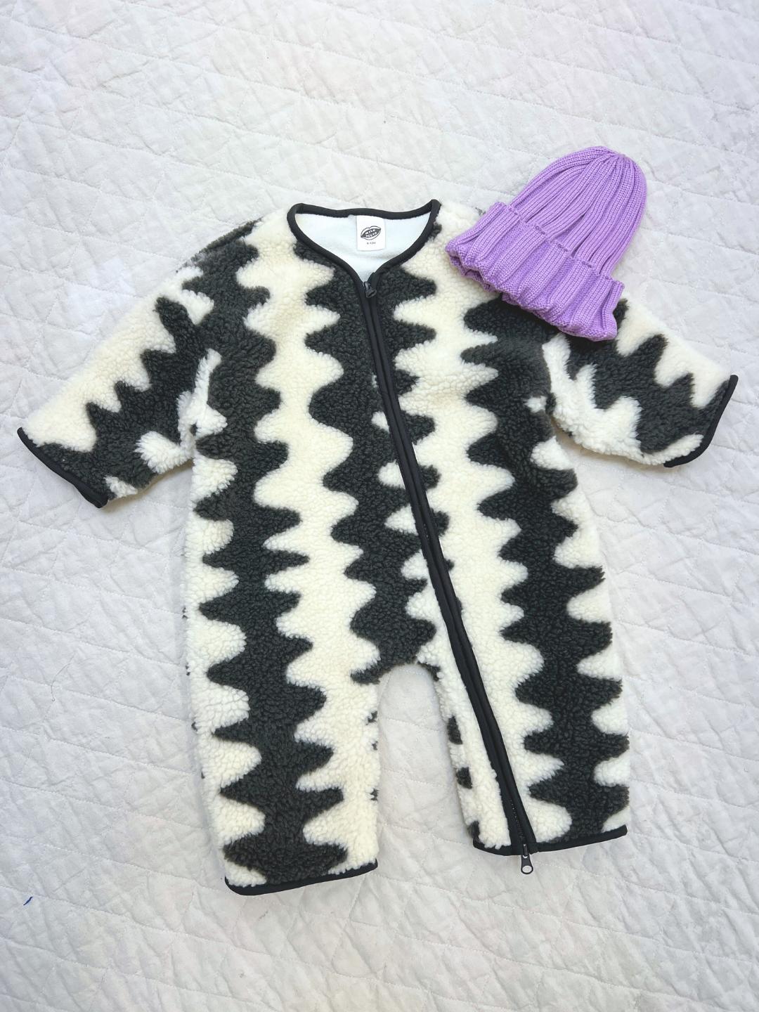 The WAVELENGTH SHERPA FLEECE STROLLER SUIT, featuring a black and white zigzag pattern and front zipper, is shown beside a folded lavender knit beanie—an ideal, cozy winter layer for babies.