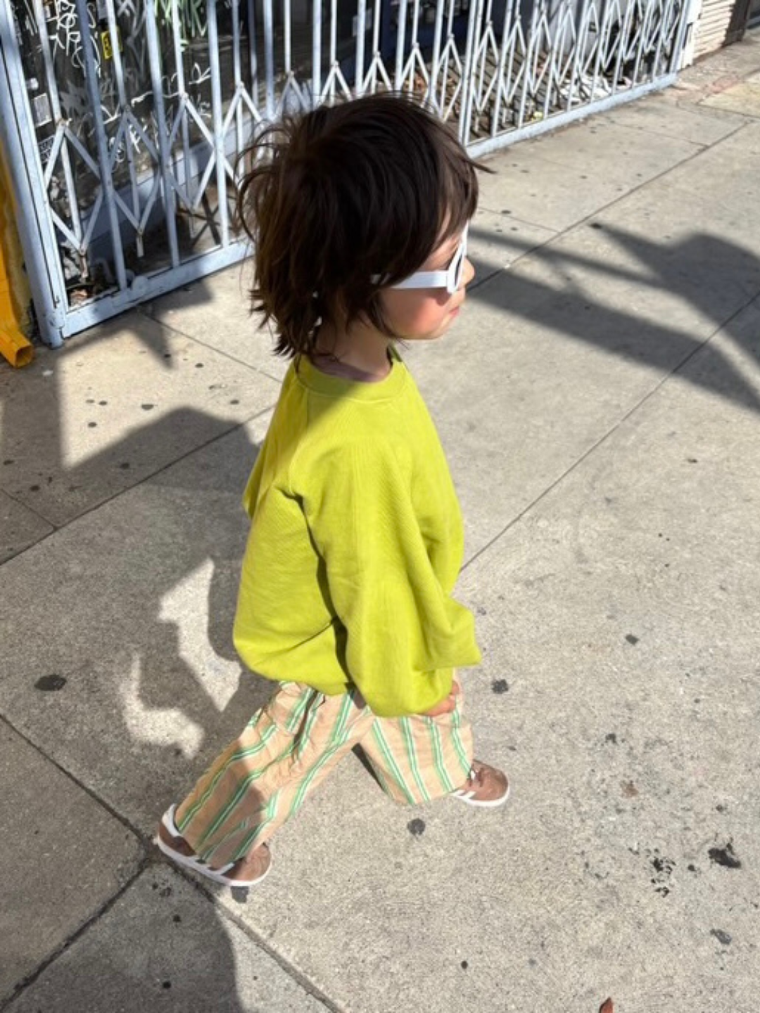 White | Child wearing a bright greenish yellow shirt and colorful pants with white rectangular sunglasses walking on a sidewalk.