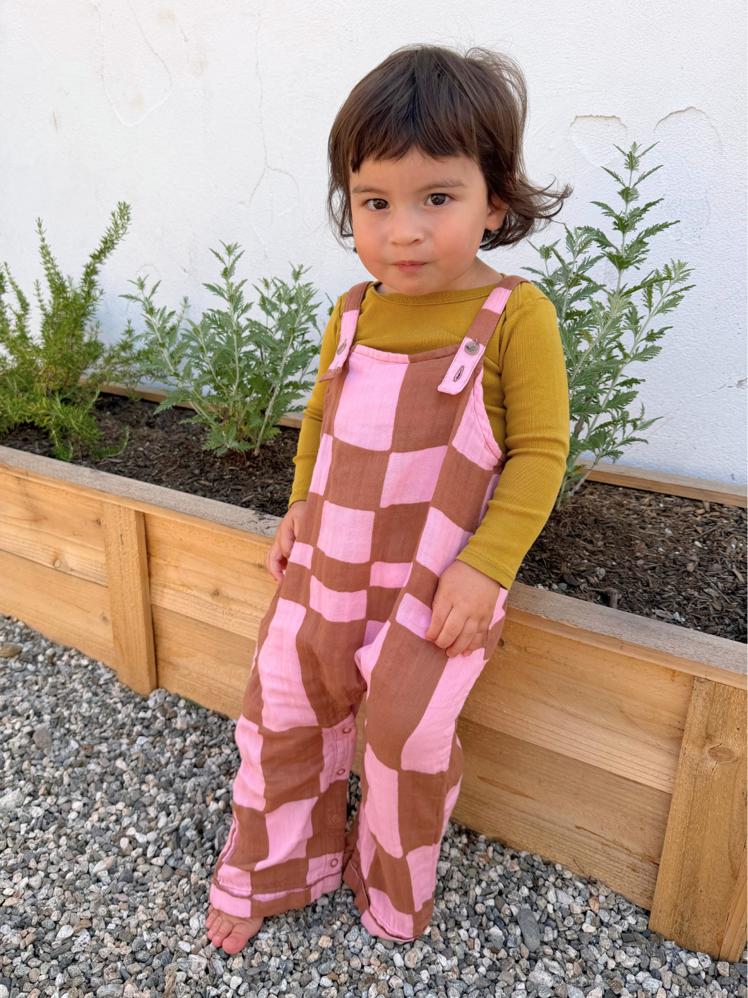 Pink Checkers | A young child stands barefoot on gravel, wearing a mustard long-sleeve shirt and MUSLIN OVERALLS made from organic cotton in a pink-and-brown checkerboard pattern, in front of a wooden planter with green plants.