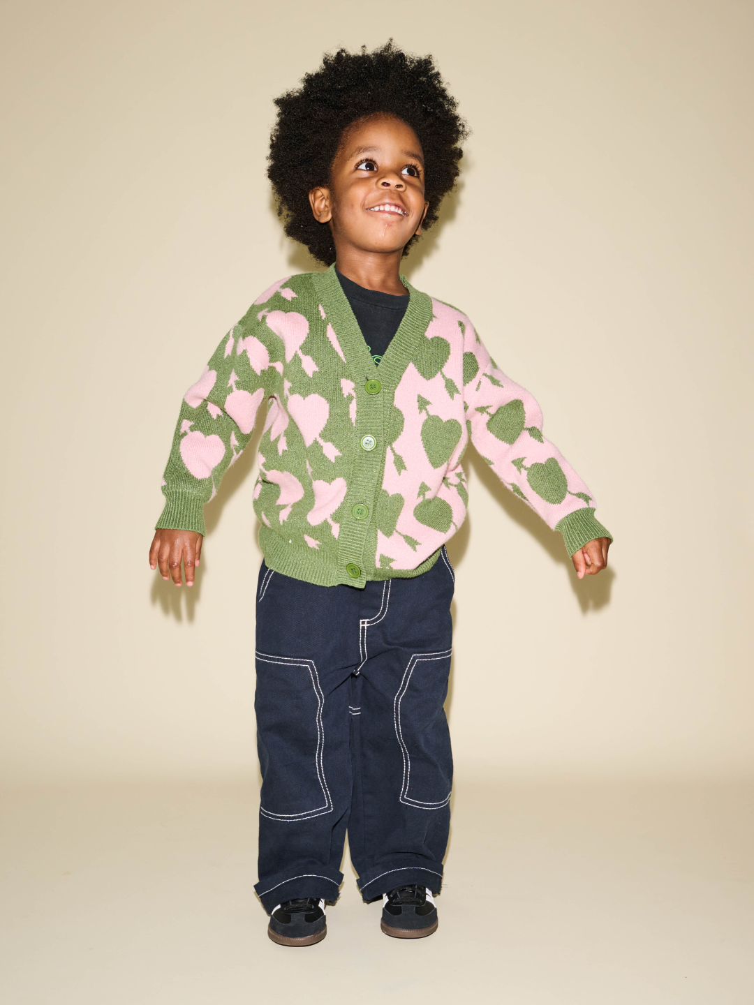 A young child with curly hair stands against a beige background, wearing the LOVESTRUCK CARDIGAN adorned with heart shapes in green and pink, paired with a black shirt, dark blue pants featuring white stitching, and black shoes. This vivid ensemble is perfect for capturing memorable photos.