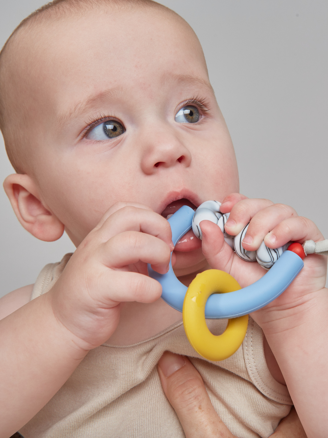 A baby in a beige tank top chews on the MT X JM ARCH RING TEETHER, a sensory-friendly US-made teether featuring blue, yellow, and white rings.