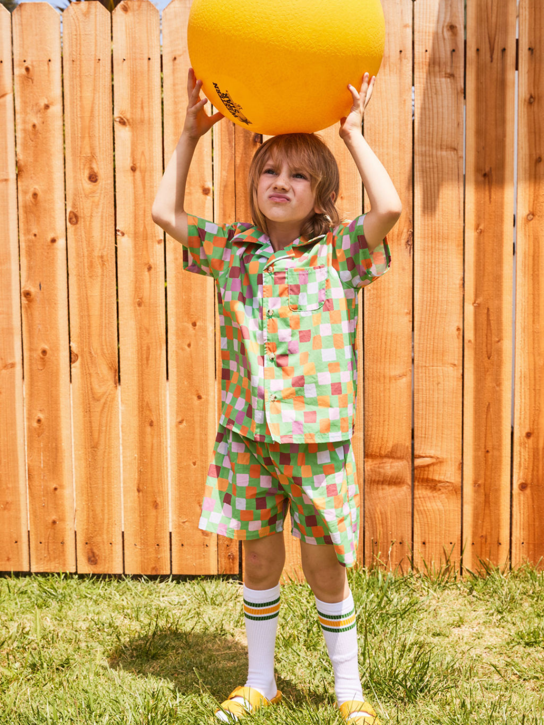 Green | A child balancing a ball on his head, wearing a kids' shirt and shorts set in a pattern of purple, pink, gold and orange squares on a green background