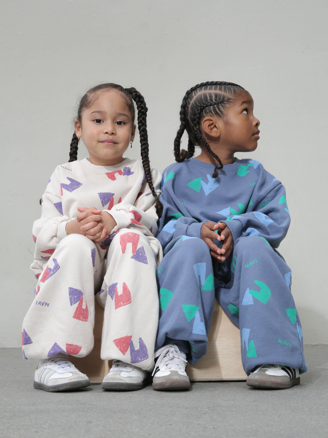 Child wearing blue sweatpants with an all over pattern of green and blue shapes and the brand name Navi, with matching crewneck sweatshirt. He sits on a wooden box against a grey background, next to another child in the same outfit in the oatmeal colorway.