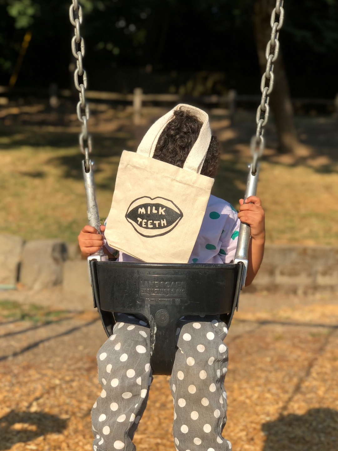 A child on swings covering their face with a mini Milk Teeth tote