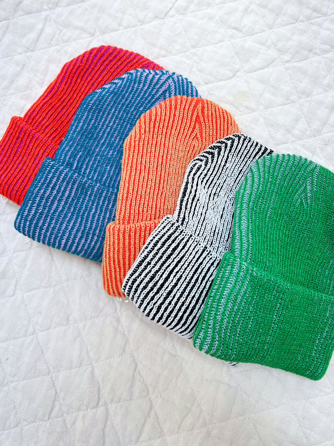 Five knit beanie hats in red, blue, orange, black and white striped, and green on a textured white background.