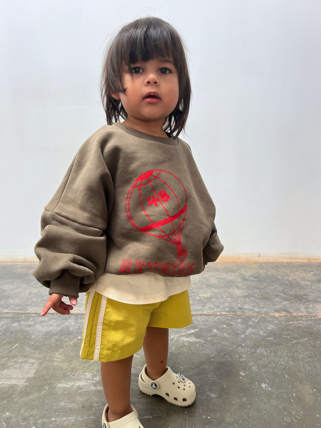 A young child with medium-length dark hair wears yellow SPRINTER SHORTS, a brown sweatshirt featuring a red balloon graphic, and white shoes, standing on a concrete floor against a plain wall.