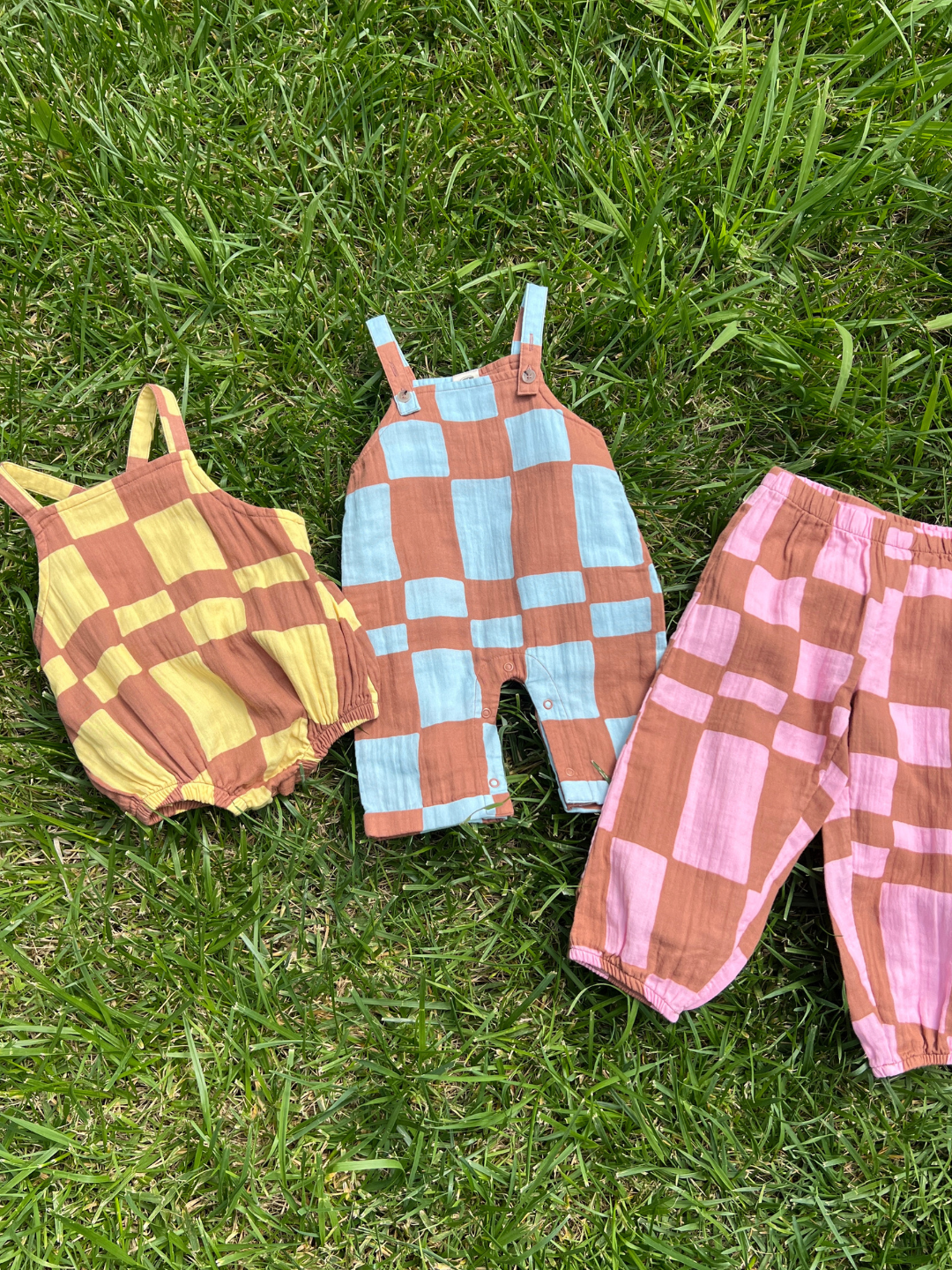 A yellow checkered romper, blue checkered baby overall and  pink checker pants, laid on grass.