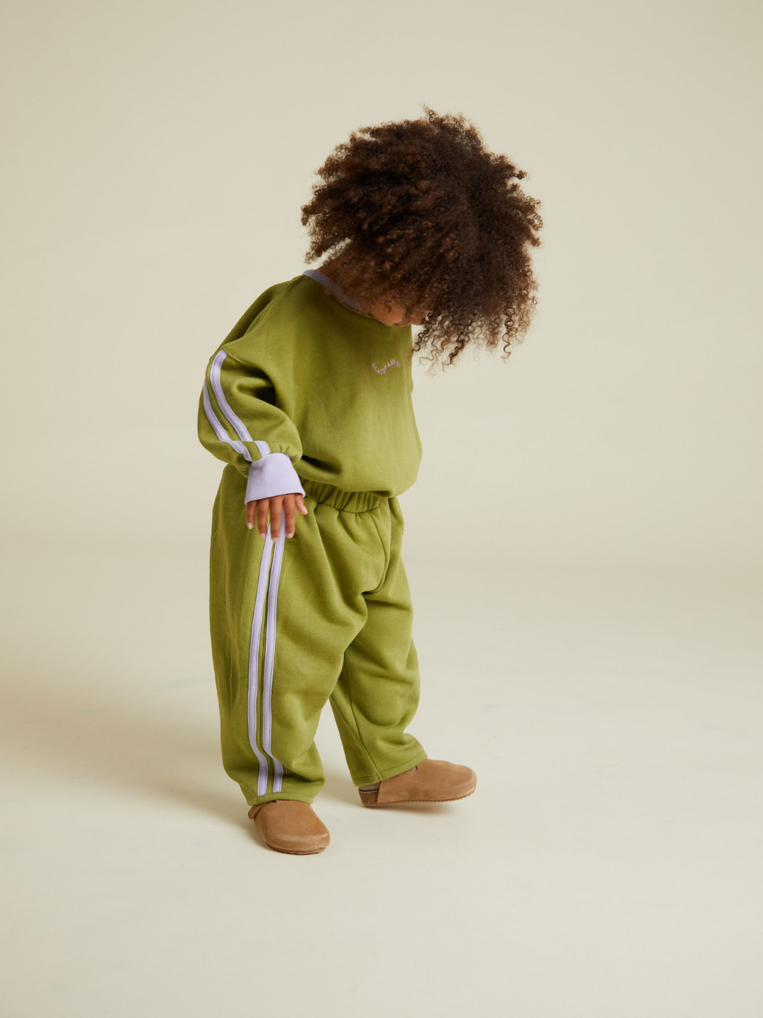 Child wearing ALVIE MATCHA SWEATPANT, featuring green fabric with white stripes and paired with brown shoes, standing on a light background and looking down.