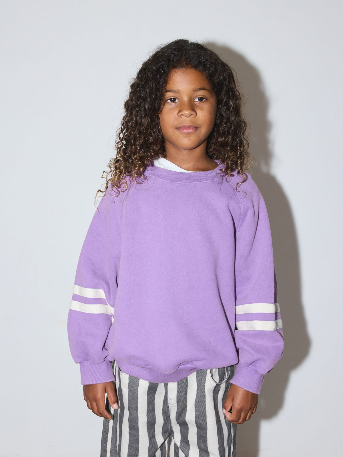Purple | Child wearing a crewneck sweatshirt in purple with two white stripes on each sleeve, with grey and white stripe pants standing in front of a white wall.