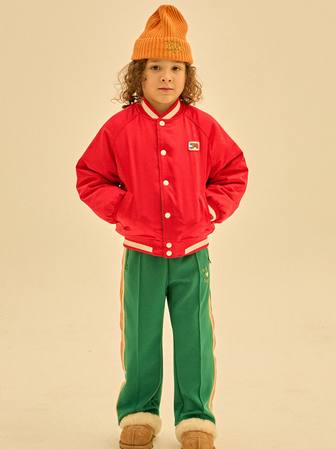 A young child with curly hair wearing an orange beanie, the BOOM REVERSIBLE JACKET in red, green pants with side stripes, and brown slippers stands with hands in pockets against a plain background. The outfit has a sporty and cozy look, perfect for merchandising alt text to highlight unique style and comfort.