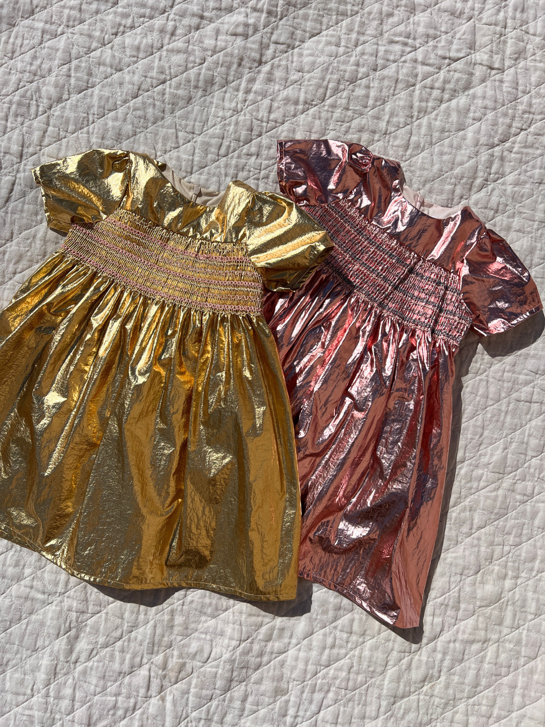 Two kids metallic party dresses, one pink, one gold, lying on a white canvas quilt. They have short sleeves, full skirts and smocking across the chest.