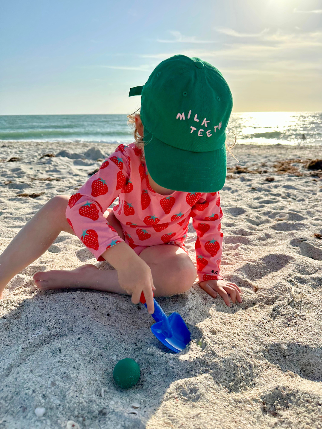 Toddler wearing the Milk Teeth baseball cap in kelly green with pink Milk Teeth embroidery, at the beach in a pink swimsuit with red strawberries.