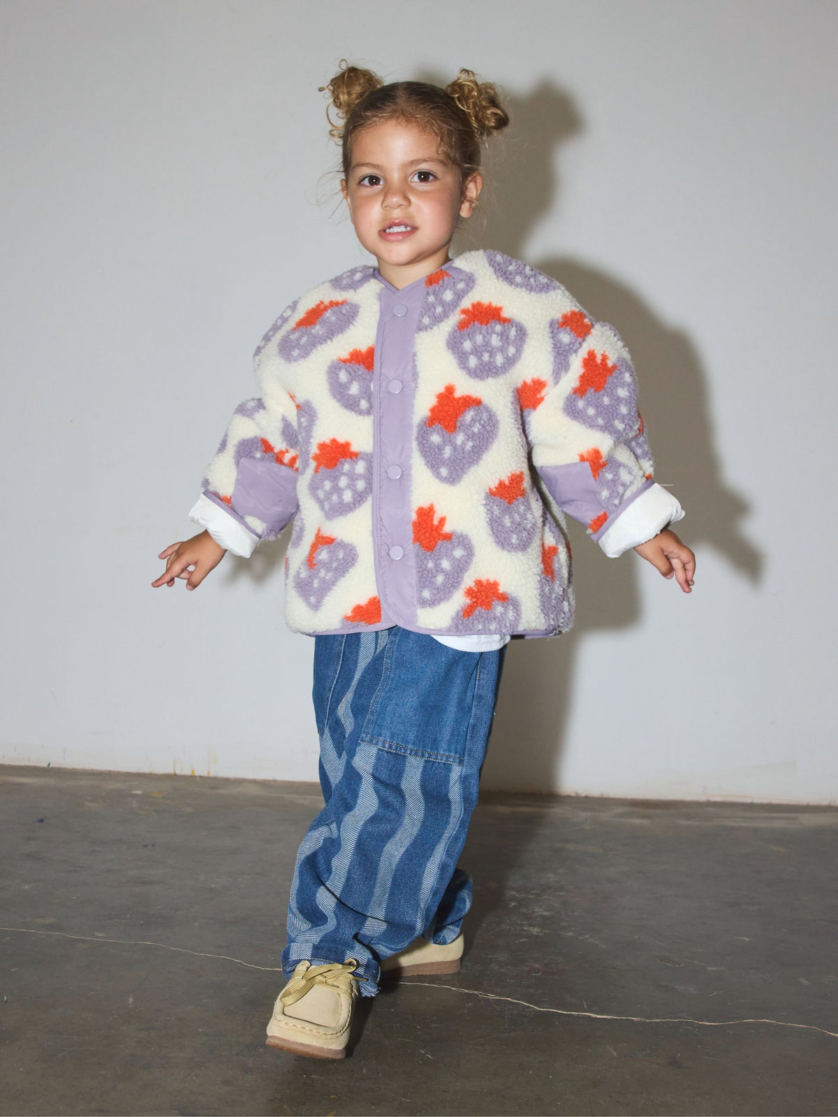 Child wearing sherpa jacket in cream fleece patterned all over in a purple and red strawberry print, with front snap closure, paired with blue striped jeans and tan shoes, standing on a concrete floor in front of a white wall