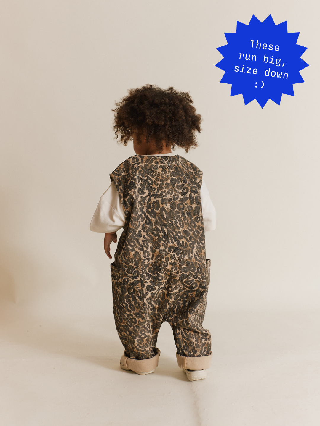 A curly-haired toddler in WREN LEOPARD OVERSIZED OVERALLs, made from organic cotton, and white shoes stands facing away in a neutral-toned room. A blue sticker calls out that these overalls run big, and to size down.