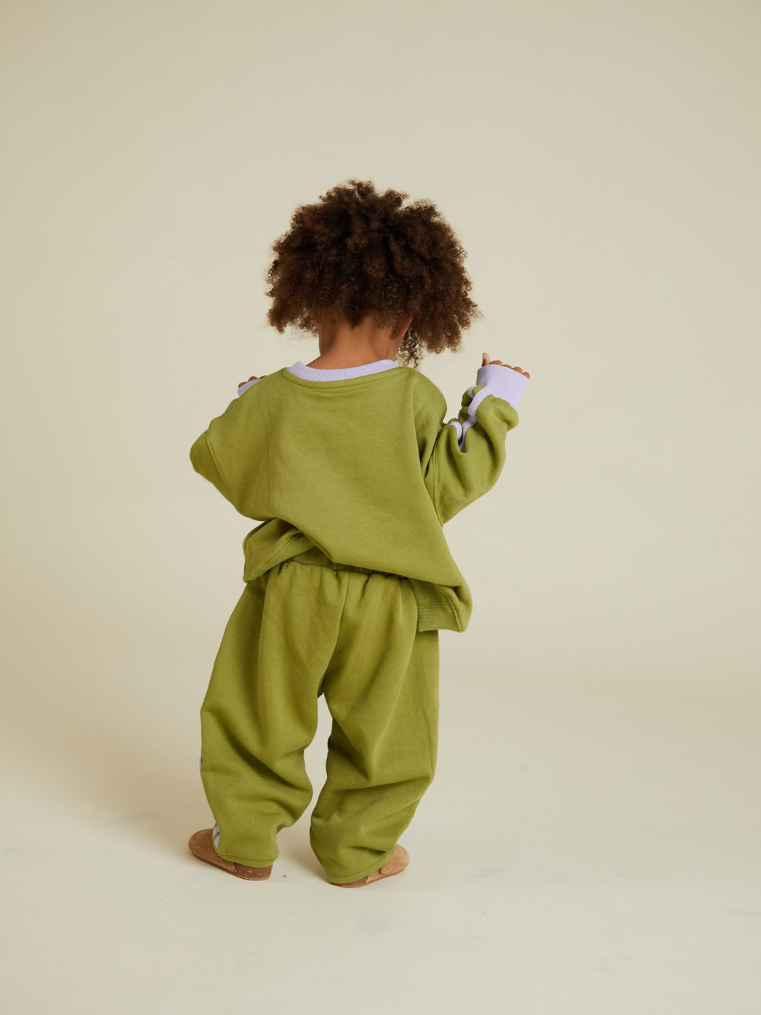 A young child with curly hair faces away, wearing ALVIE MATCHA SWEATPANT and a matching oversized sweatshirt against a plain beige background.