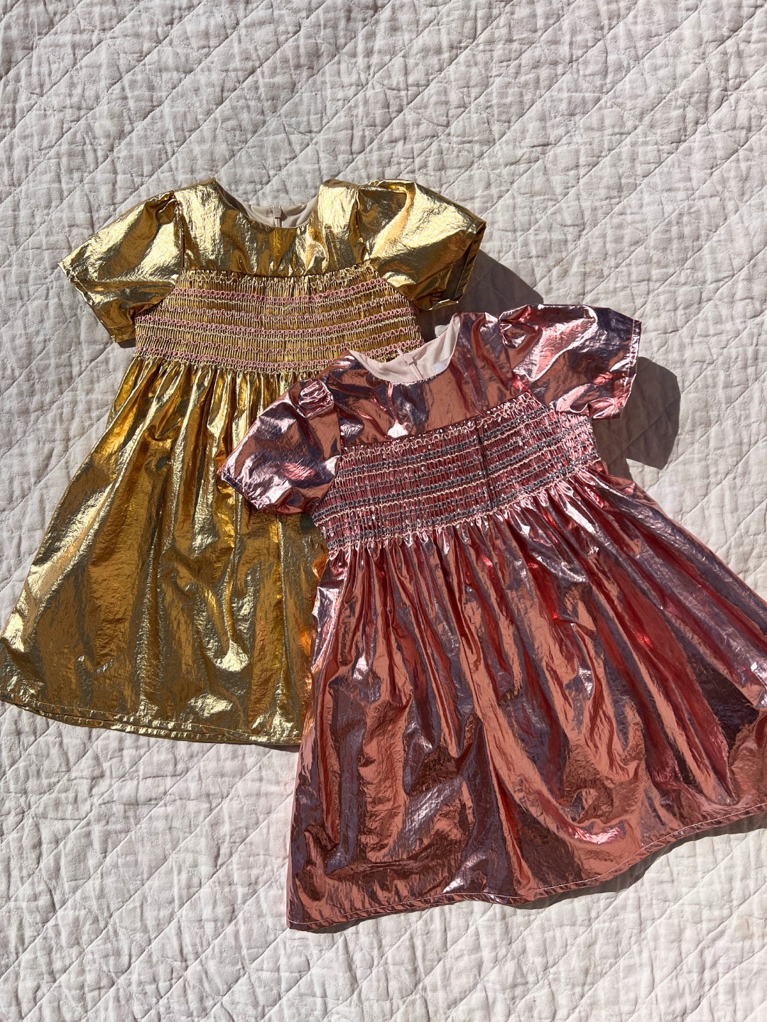 Rose | Two kids metallic party dresses, one pink, one gold, lying on a white canvas quilt. They have short sleeves, full skirts and smocking across the chest.