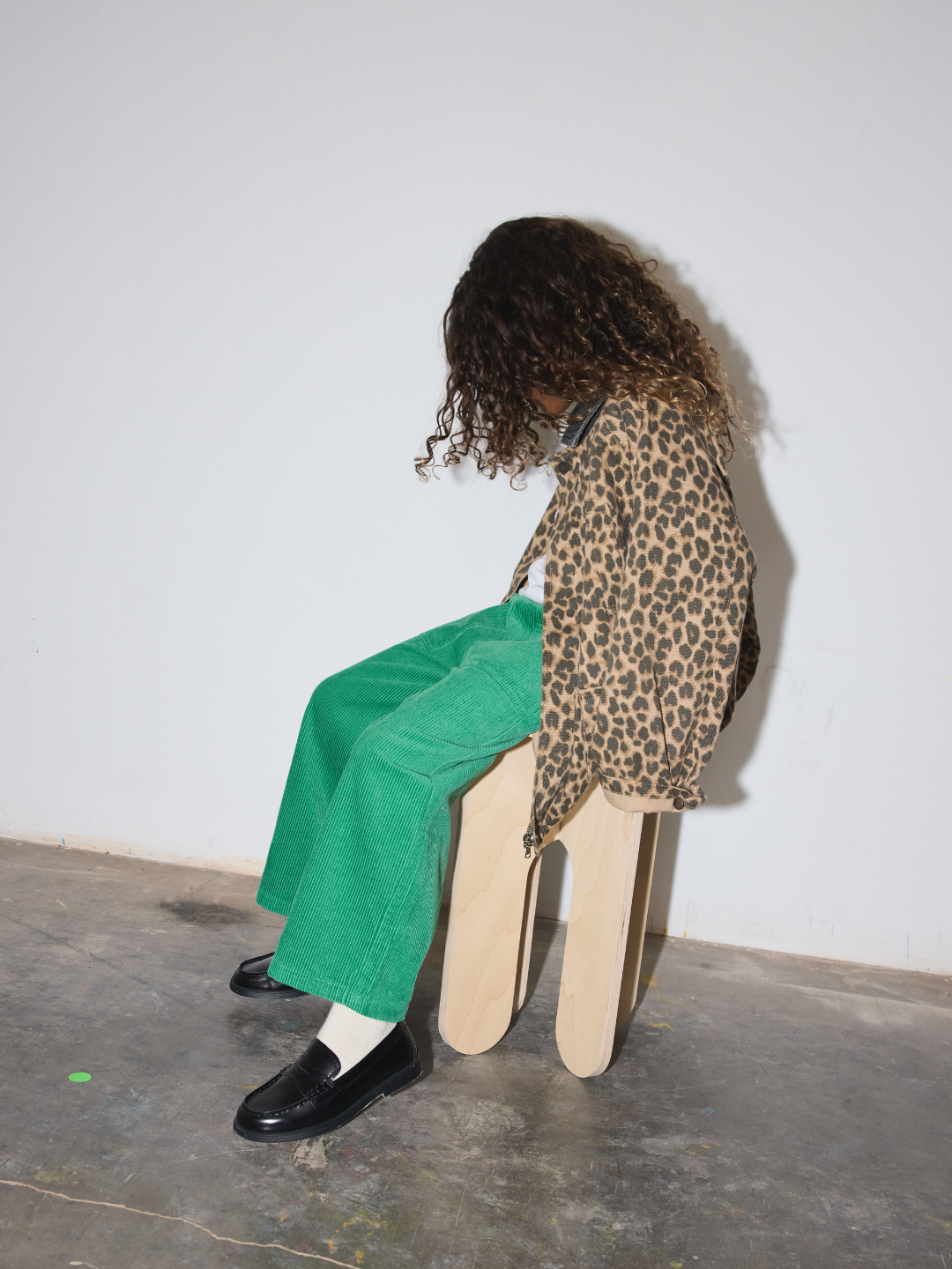  A child with curly dark hair sits on a light wood stool, wearing a leopard-print jacket, bright green corduroy pants, white socks, and black loafers against a plain white wall and concrete floor.