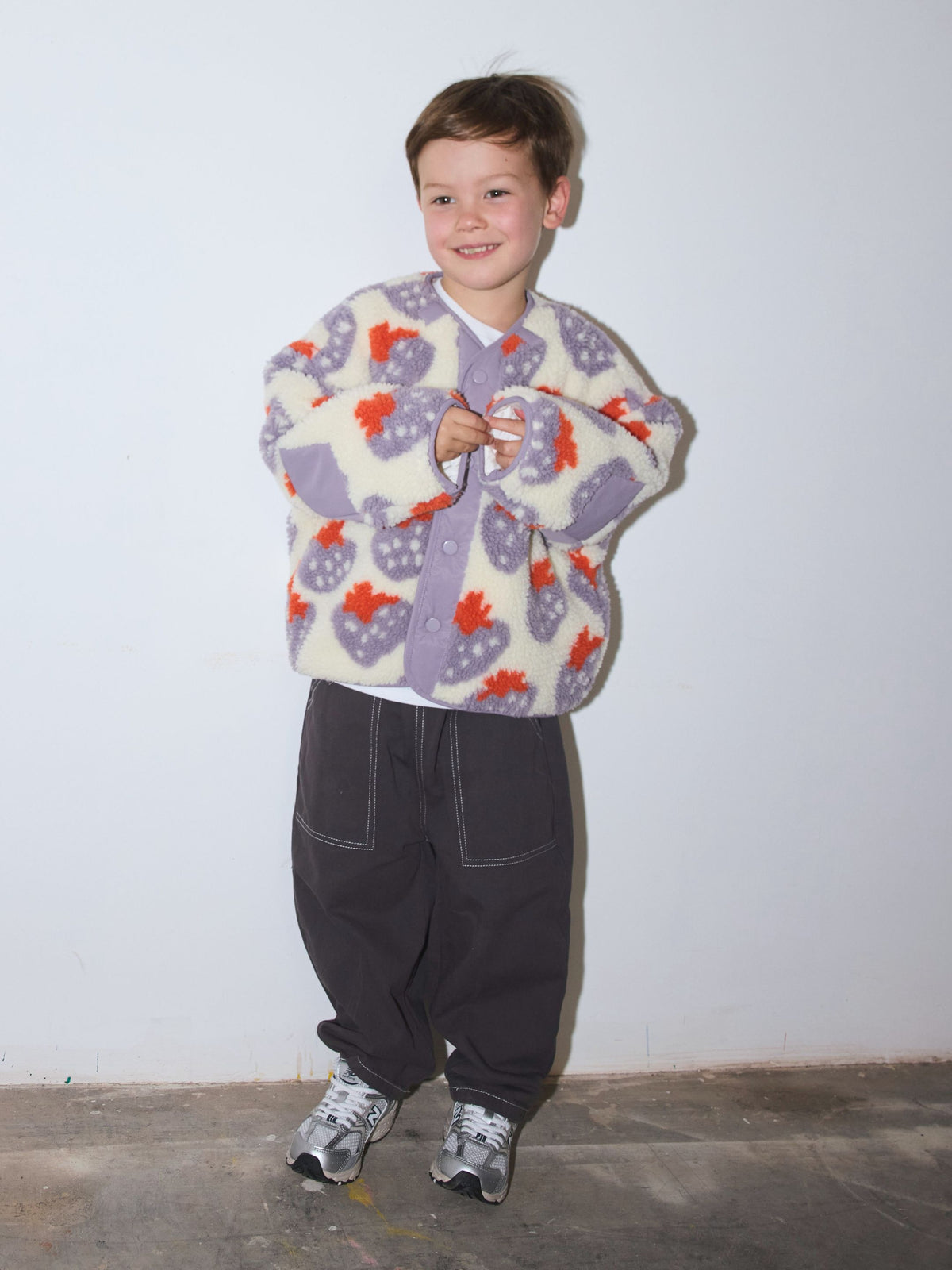 Boy wearing a sherpa jacket in cream fleece patterned all over in a purple and red strawberry print, with front snap closure, paired with grey baggy pants and grey and silver sneakers, standing on a concrete floor in front of a white wall