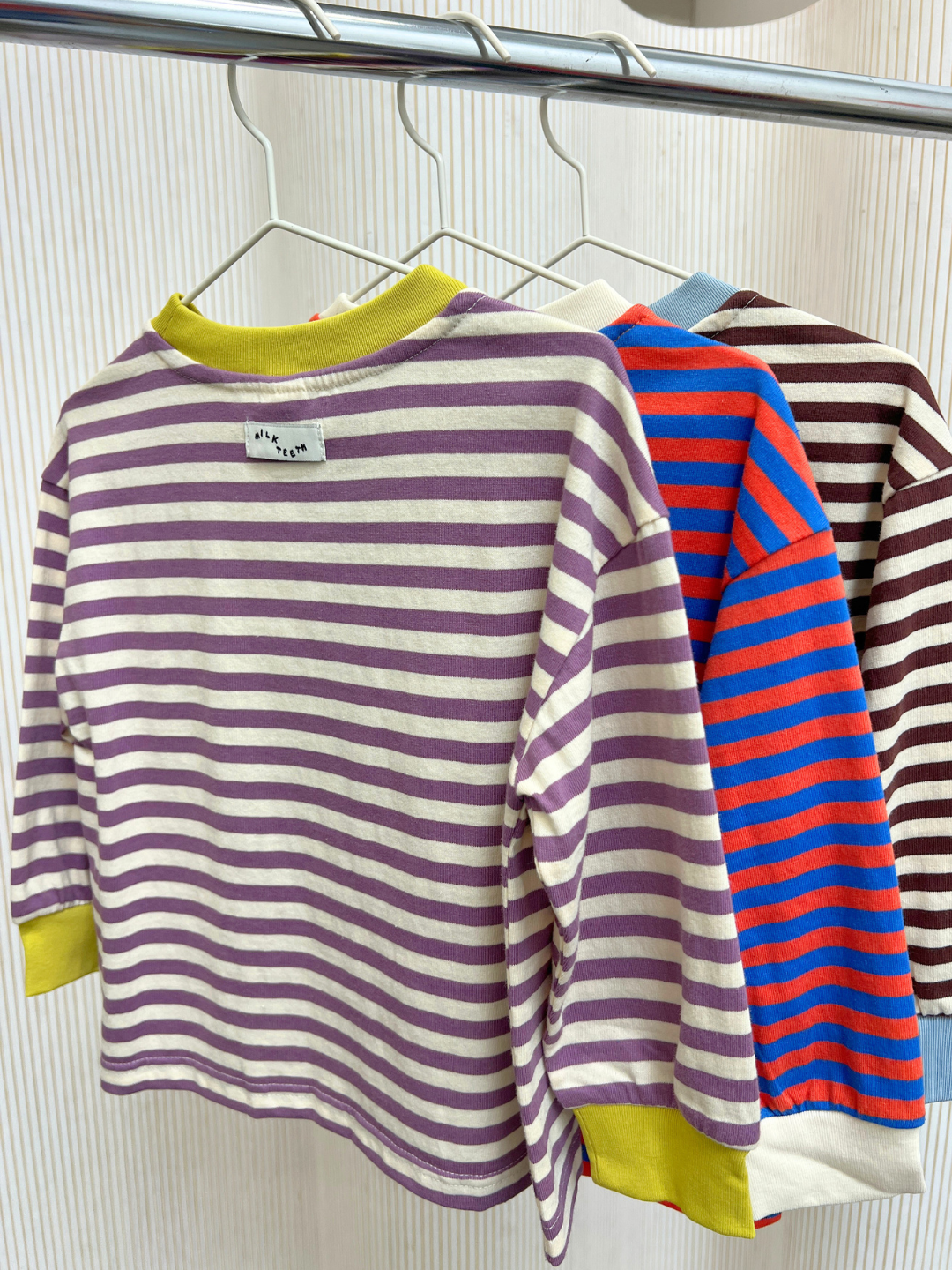 Purple/Ecru | Three STRIPE LONGSLEEVE tees, featuring a relaxed fit and crafted from 100% cotton jersey striped fabric in different color combinations, hang on white hangers against a light background.