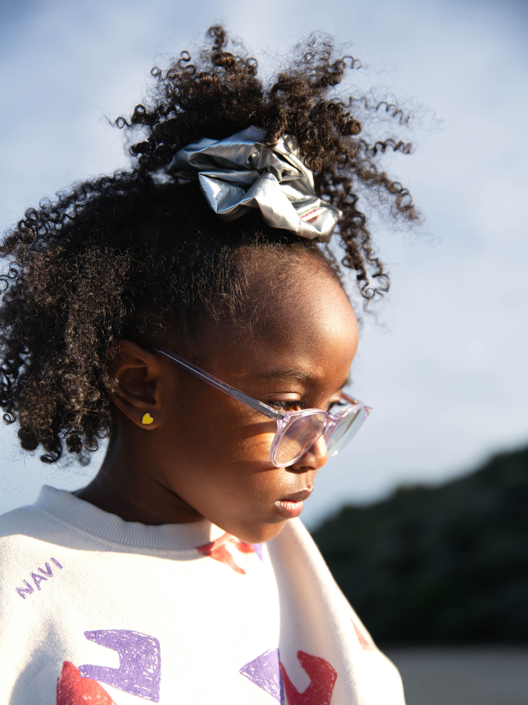 Girl wearing the stardust oversized scrunchie in metallic silver. She has a half ponytail in her curly hair and wears pink glasses, gold heart earrings and a cream sweatshirt printed with purple and red shapes. She is outdoors.