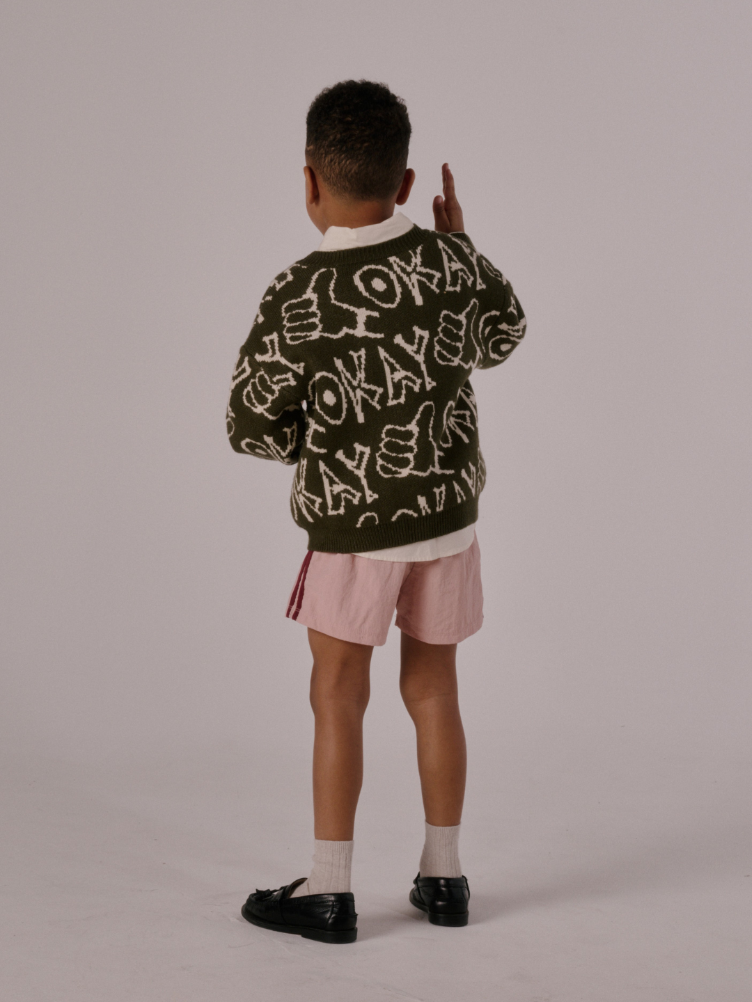A young child faces away in the OKAY SWEATER, featuring "OKAY" and thumbs-up graphics, paired with pink shorts, white socks, and black shoes against a plain background.