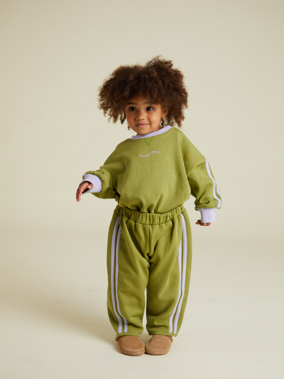 A young child with curly hair wears the ALVIE MATCHA SWEATPANT, part of a green matching set with white stripes and tan shoes, standing against a plain light background.
